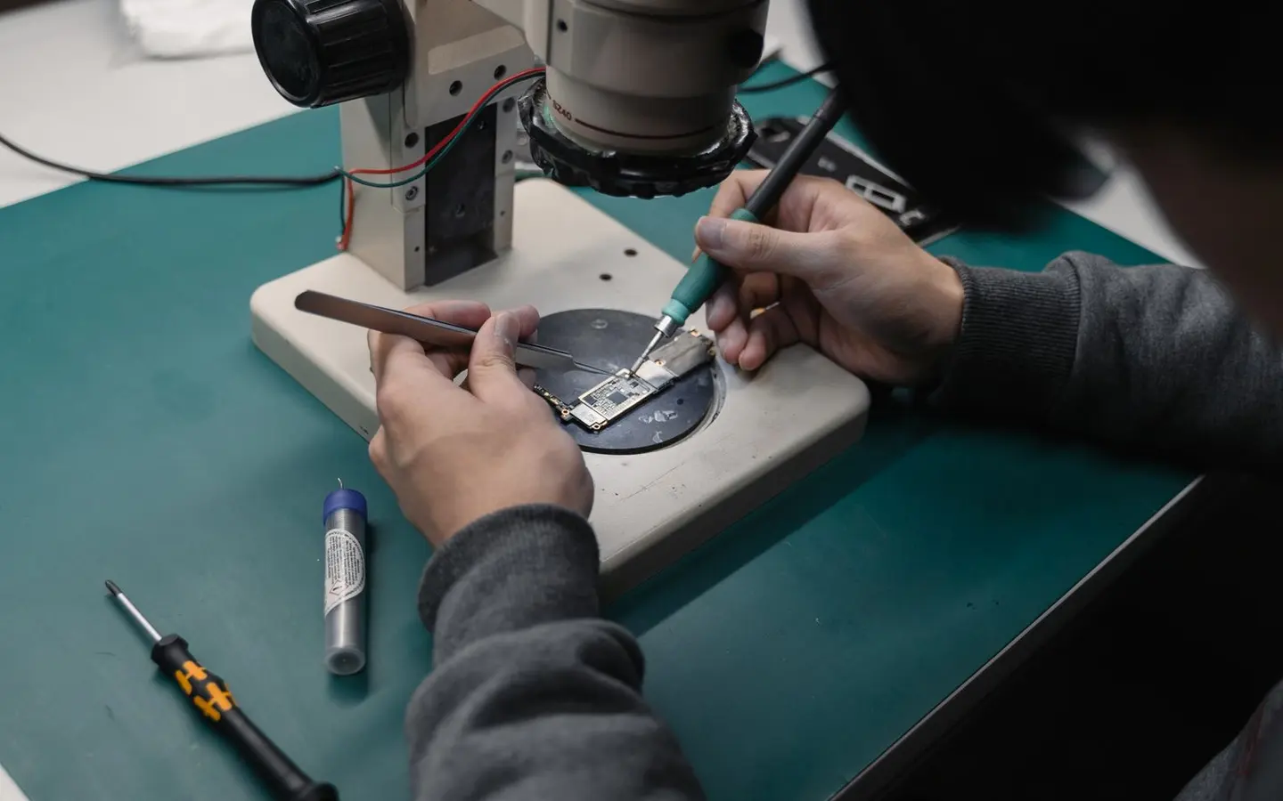 Mains tenant une pince et un fer a souder fin sur une carte mere, sous l'objectif du microscope