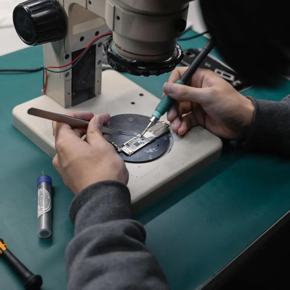 Mains tenant une pince et un fer a souder fin sur une carte mere, sous l'objectif du microscope